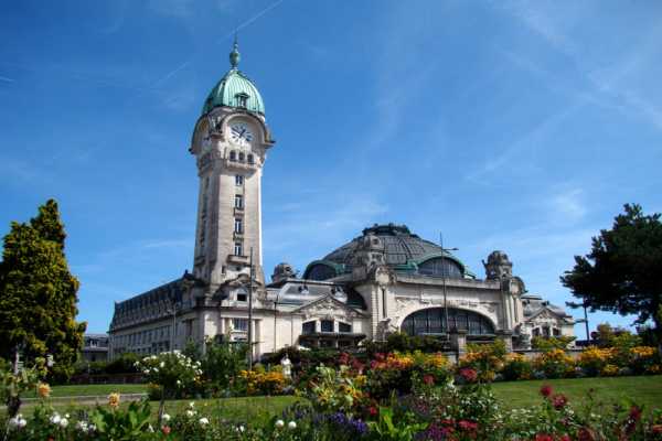 Ancient Building With A Tower And Clock On Top, Beuatiful Garden Of Colorful Flowers In The Frontyard 